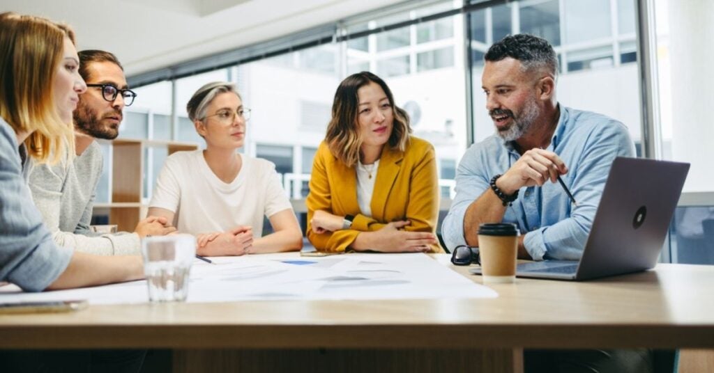 Four people sitting around a table listening to a fifth man talking