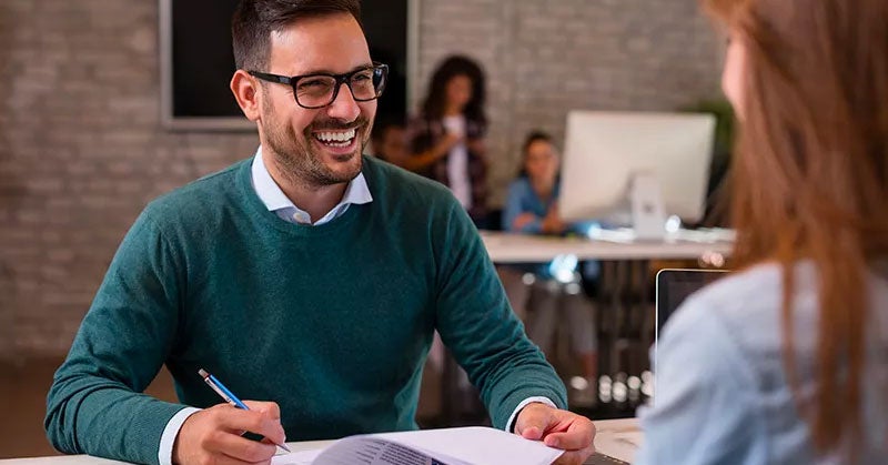 Man in a sweater chatting across a desk with a second person