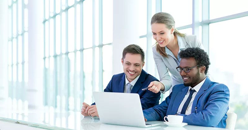 Three people in suits looking at a laptop in a glass office