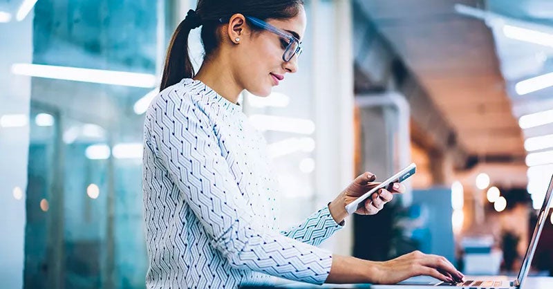 Woman at a laptop reading messages on a smartphone