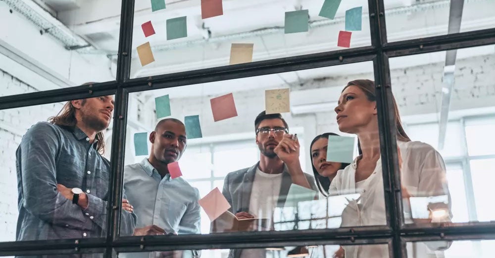 Four people looking at a glass wall covered in post-it notes