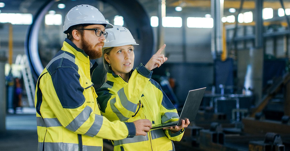 Two people wearing hardhats and yellow safety jackets looking at machinery