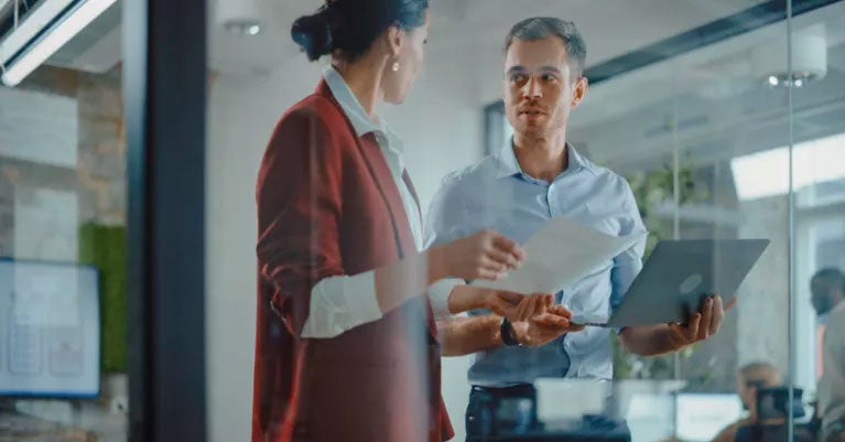 Two people holding a document and a laptop in a glass office