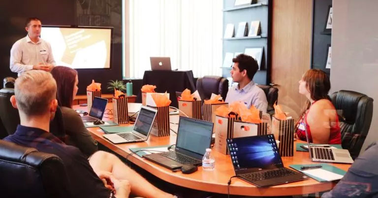 People sitting at a long table with laptops and a TV screen