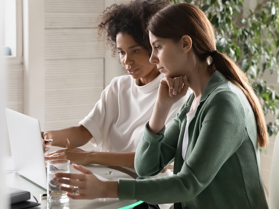 Confident African American mentor teaching new employee, using laptop, pointing on laptop screen, helping with corporate software, diverse colleagues working on project, sharing ideas, talking