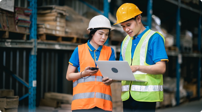 Two people wearing hardhats in a warehouse checking data on a laptop