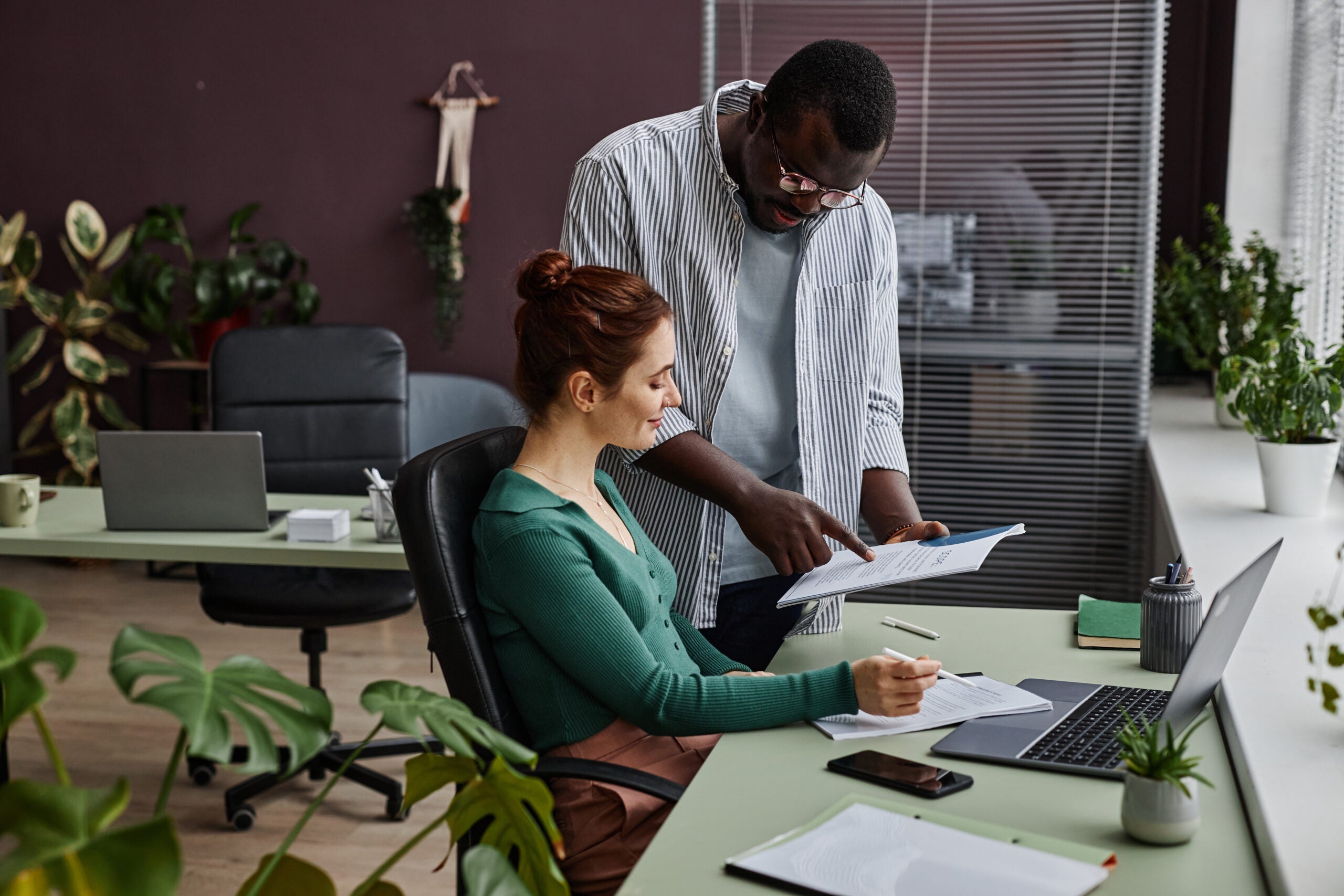 Side view portrait of two people discussing project and reading documents at workplace in office decorated with plants