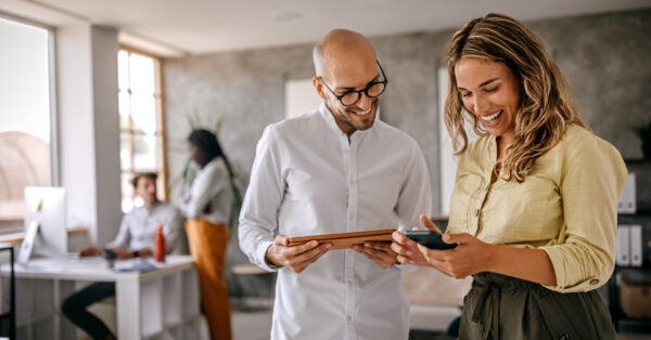 Cheerful and smiling young successful female businesswoman standing with colleague looking at smartphone in modern office and coworking space