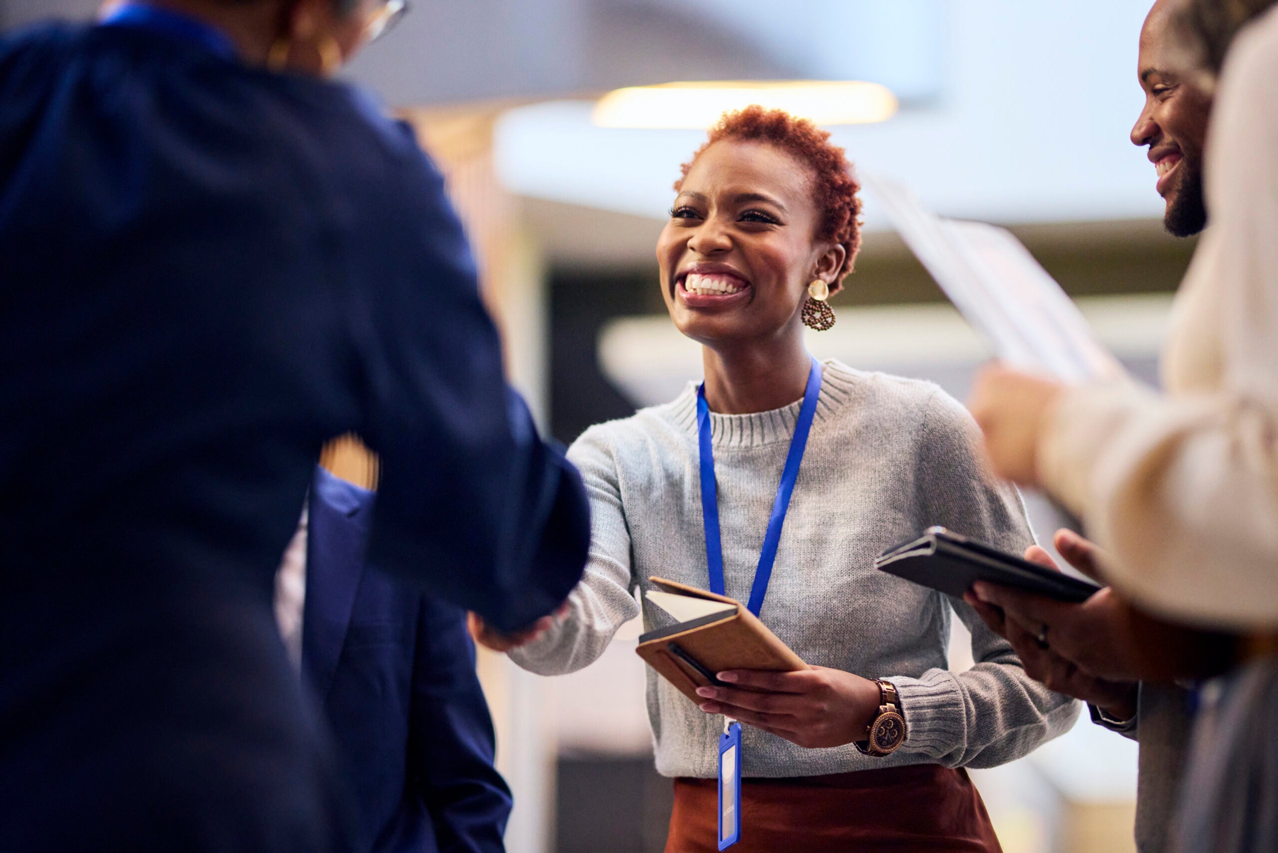 A vibrant businesswoman with short red hair enthusiastically shakes hands at a conference. She wears a grey sweater and brown skirt, holding a notebook. Other attendees are partially visible in the background.