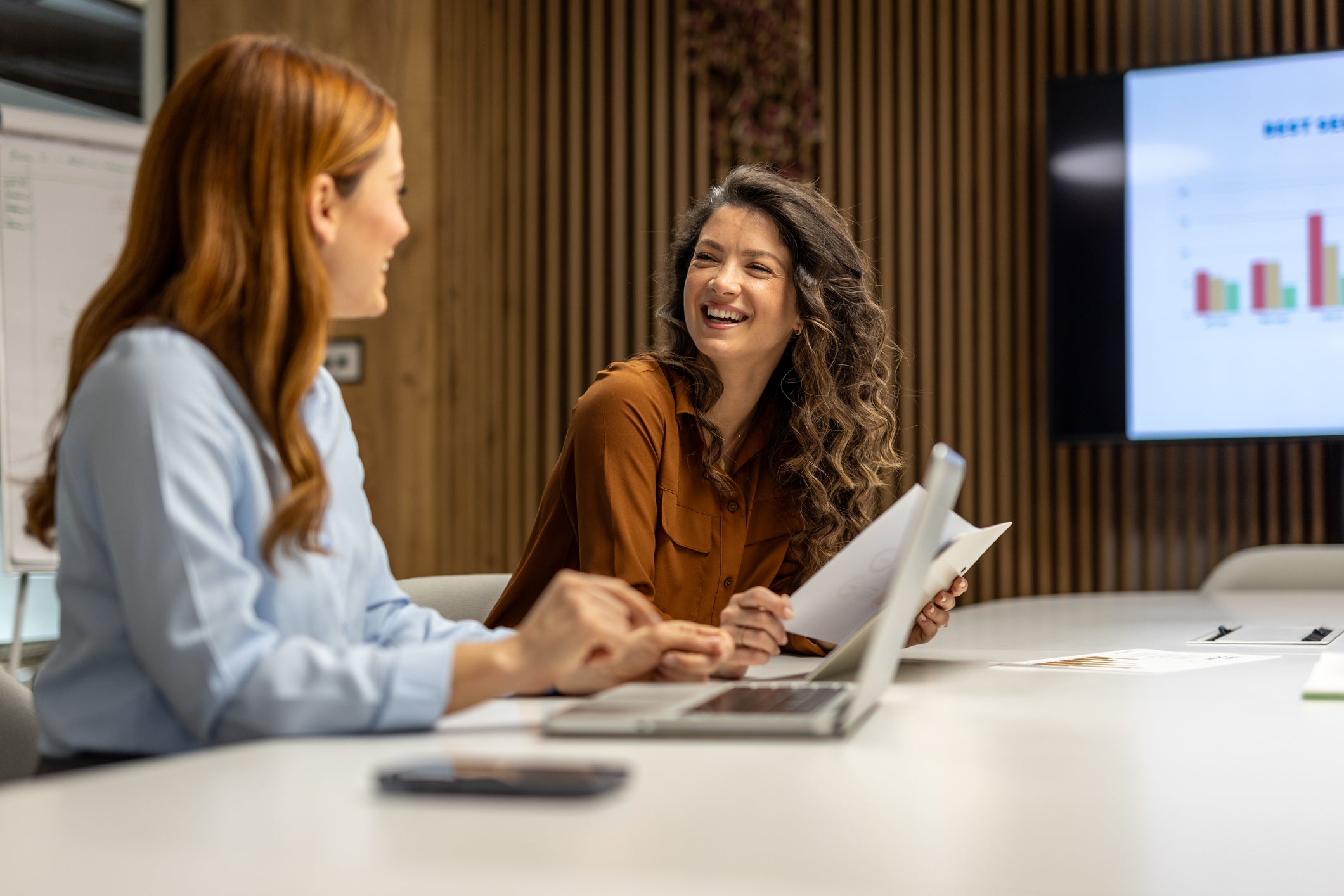 Two businesswomen are sitting at a table in a modern office, collaborating on a project, sharing documents, using a laptop, and smiling, with a graph displayed on a screen in the background
